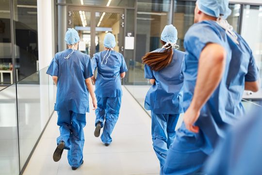 Doctors in blue surgical gowns rush down a hallway; shot from behind