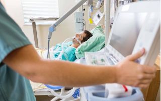 Beatmeter Patient im Krankenbett, Pflegekraft im Vordergrund bedient ein medizinisches Gerät