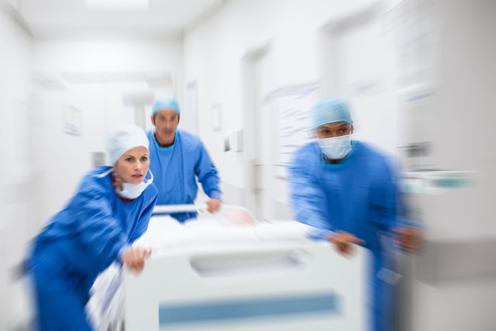 Doctors in blue surgical gowns rush down the hallway with a hospital bed.