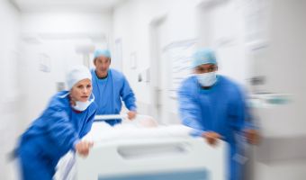 Doctors in blue surgical gowns rush down the hallway with a hospital bed.