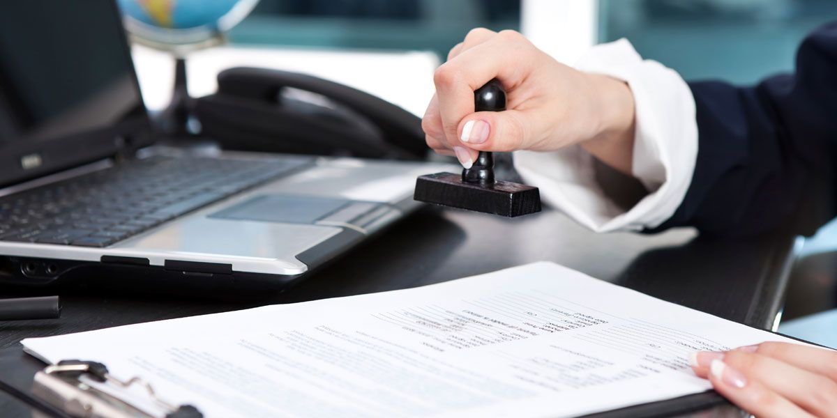 The hand of a person outside the frame stamps a document lying on a desk