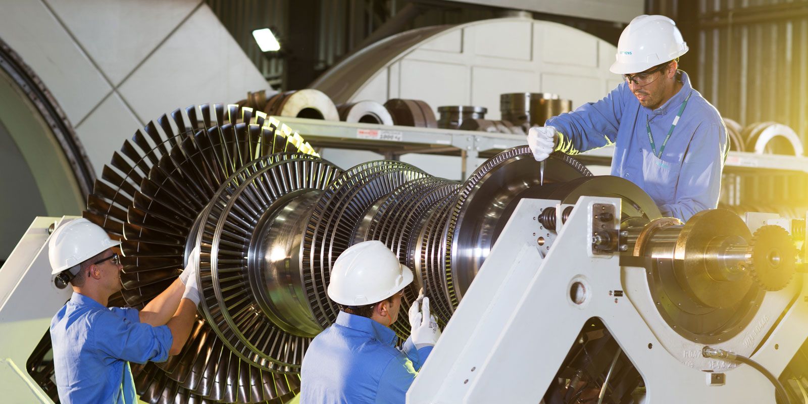 Three engineers in blue overalls and white hard hats work on an engine