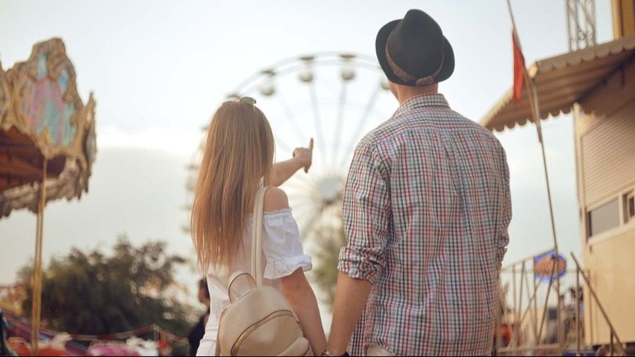 A couple stands with their backs to the camera in an amusement park, she points to a Ferris wheel in the background